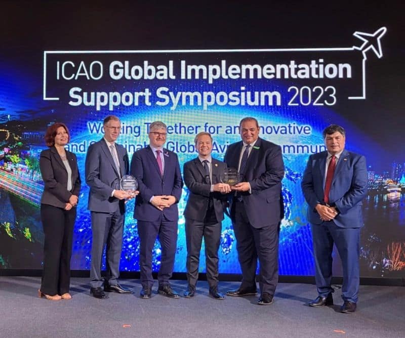Six people in business suits stand in front of a backdrop that reads ICAO Global Implementation Support Symposium 2023.